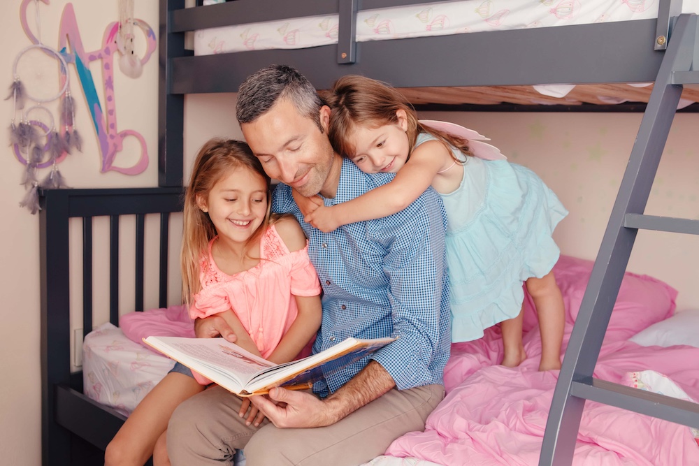 Dad reading book to daughters girls. Family of three people sitting on bed in bedroom