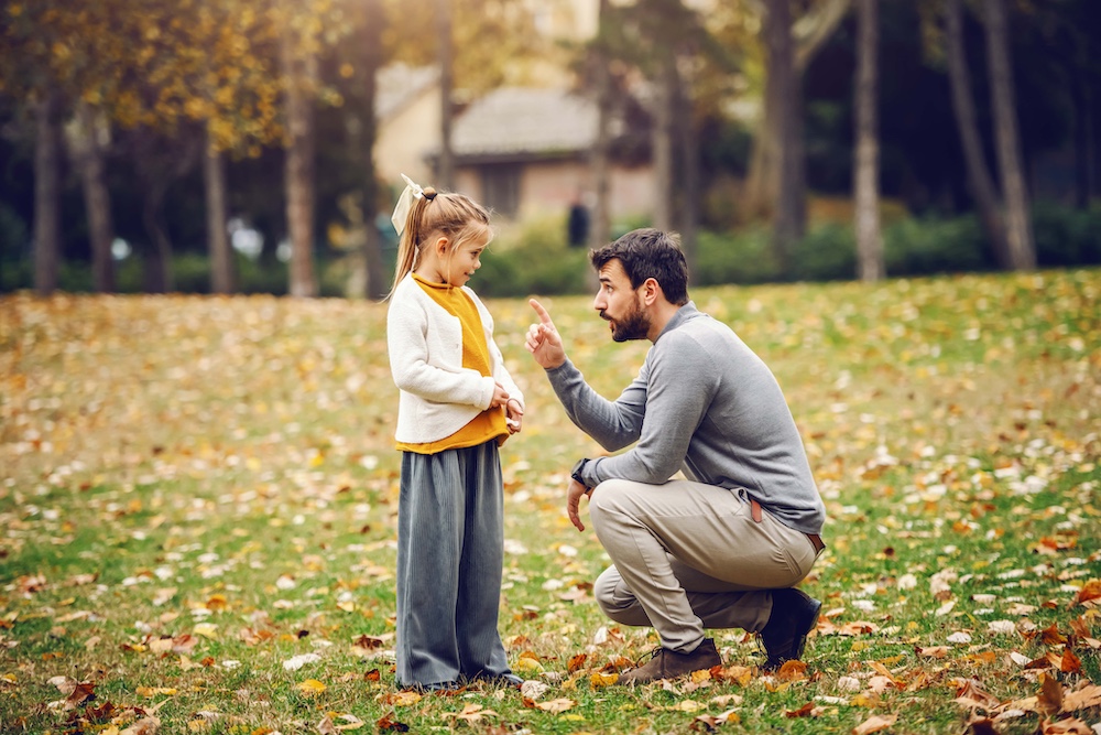 Devilish cute little girl with ponytail standing and looking at her father. Father crouching and reprimanding her. Autumn in park